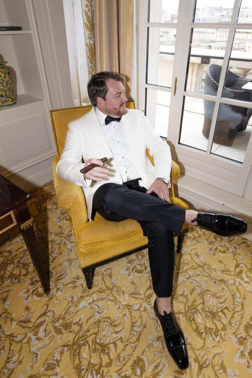 Groom in a white tuxedo relaxing with a cigar in a luxury Paris hotel suite, captured by Olivier Neuville Photography.